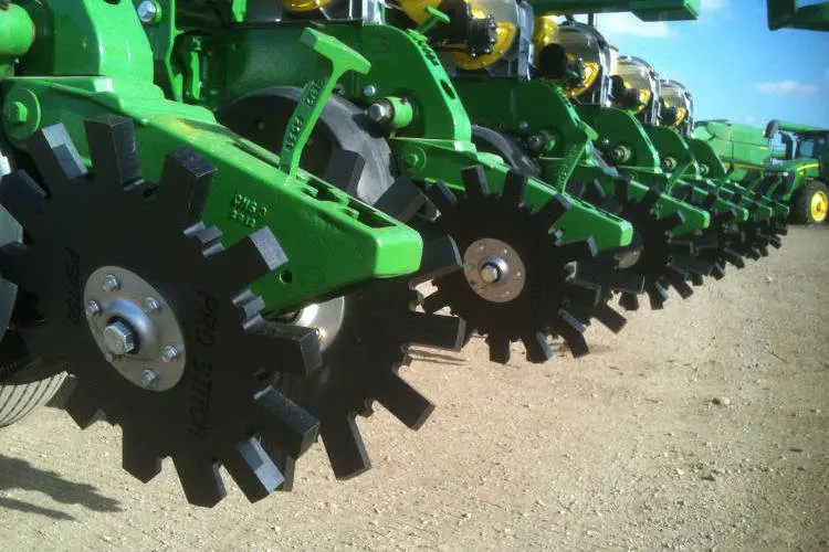 Close-up of green agricultural planter equipment with black notched closing wheels on a dirt surface under a clear blue sky.